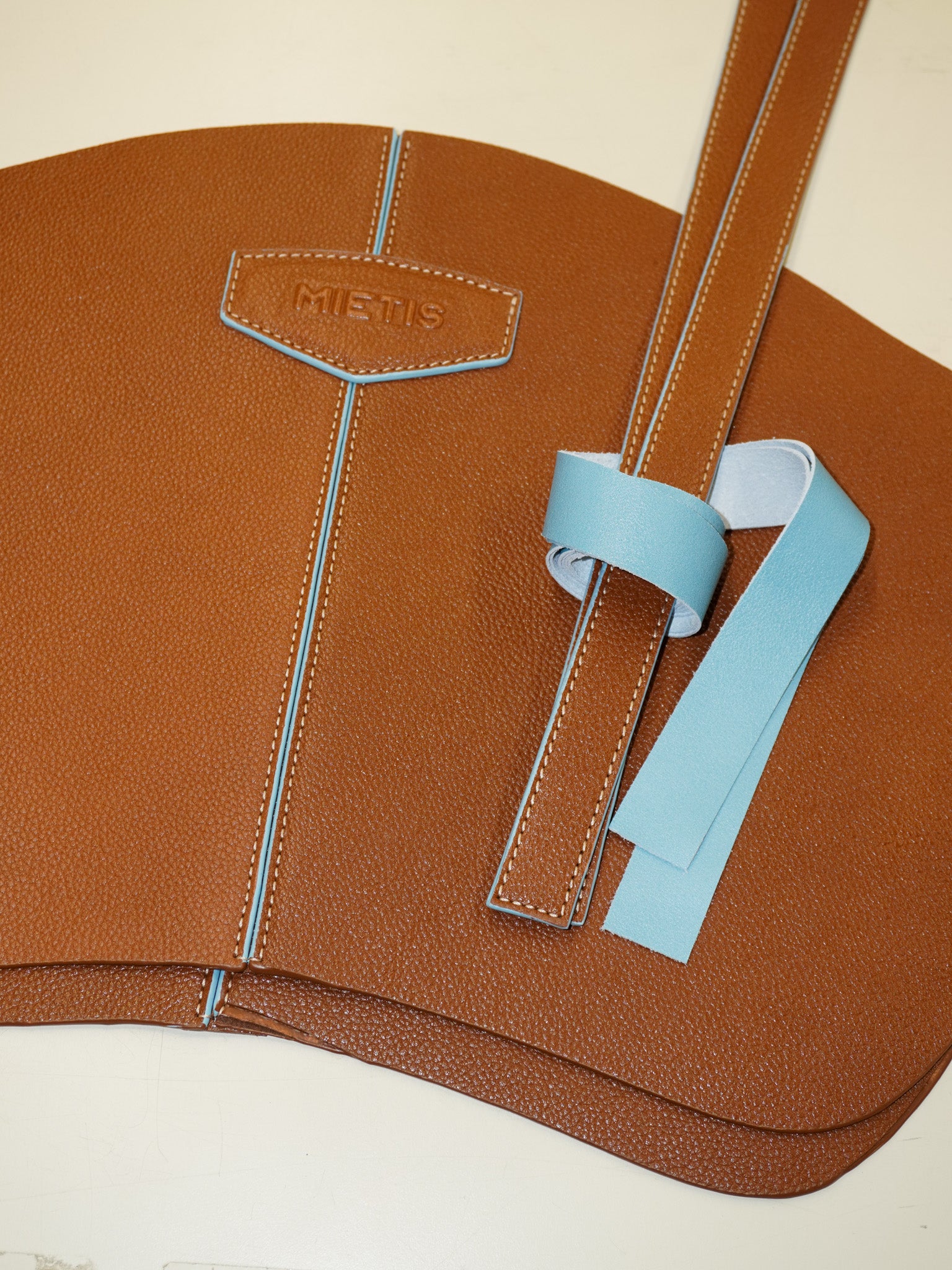 Brown leather bag with blue accents and 'Mietis' branding on a beige background
